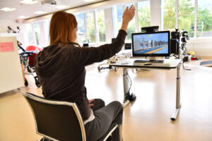 A woman sat in a chair lifting her arm in front of a screen to operate a game